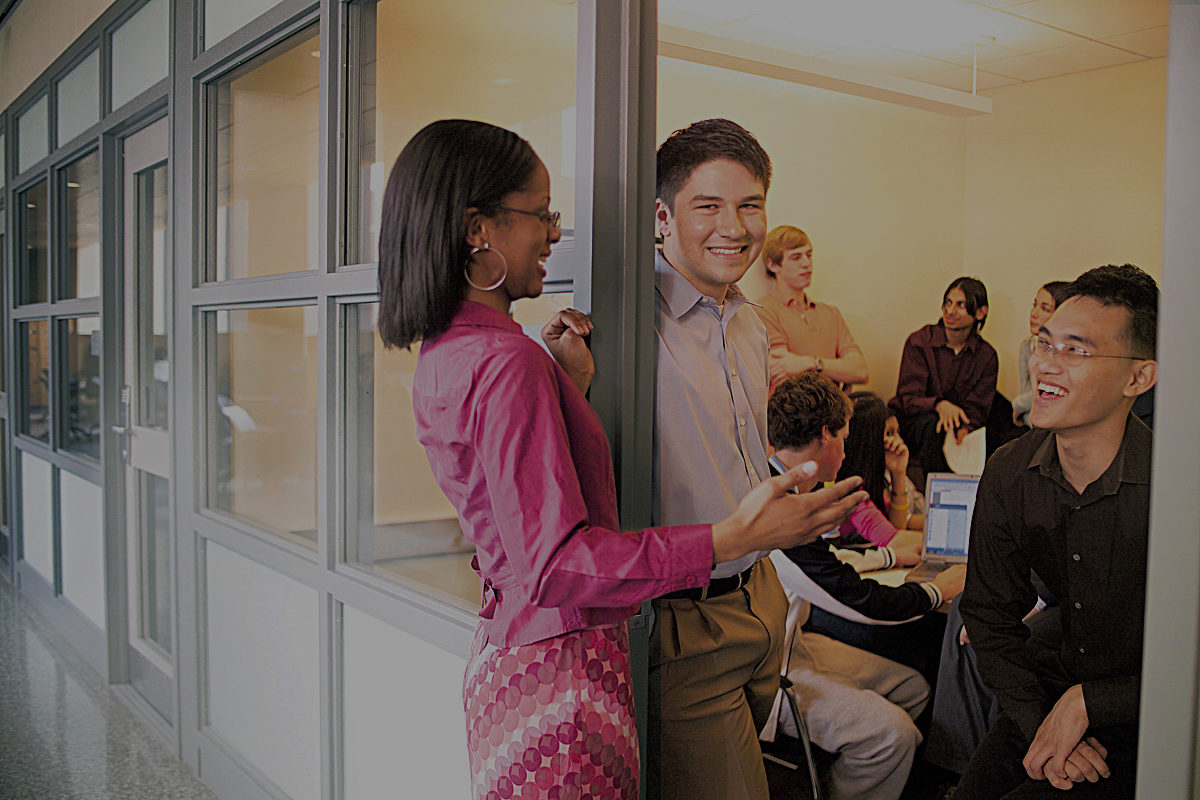 A small group of people socializing and smiling in an office or meeting environment. Some are seated, and others are standing near a partition.